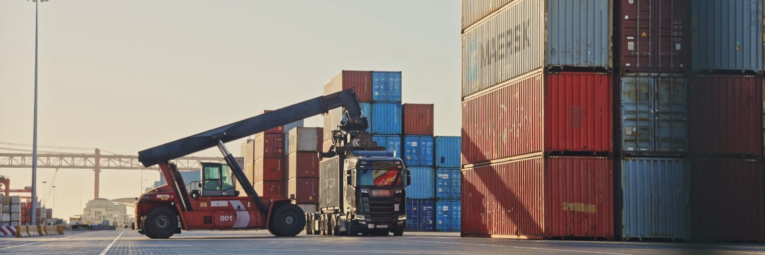 A shipping container is being unloaded from a truck at a port terminal.