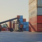 A shipping container is being unloaded from a truck at a port terminal.